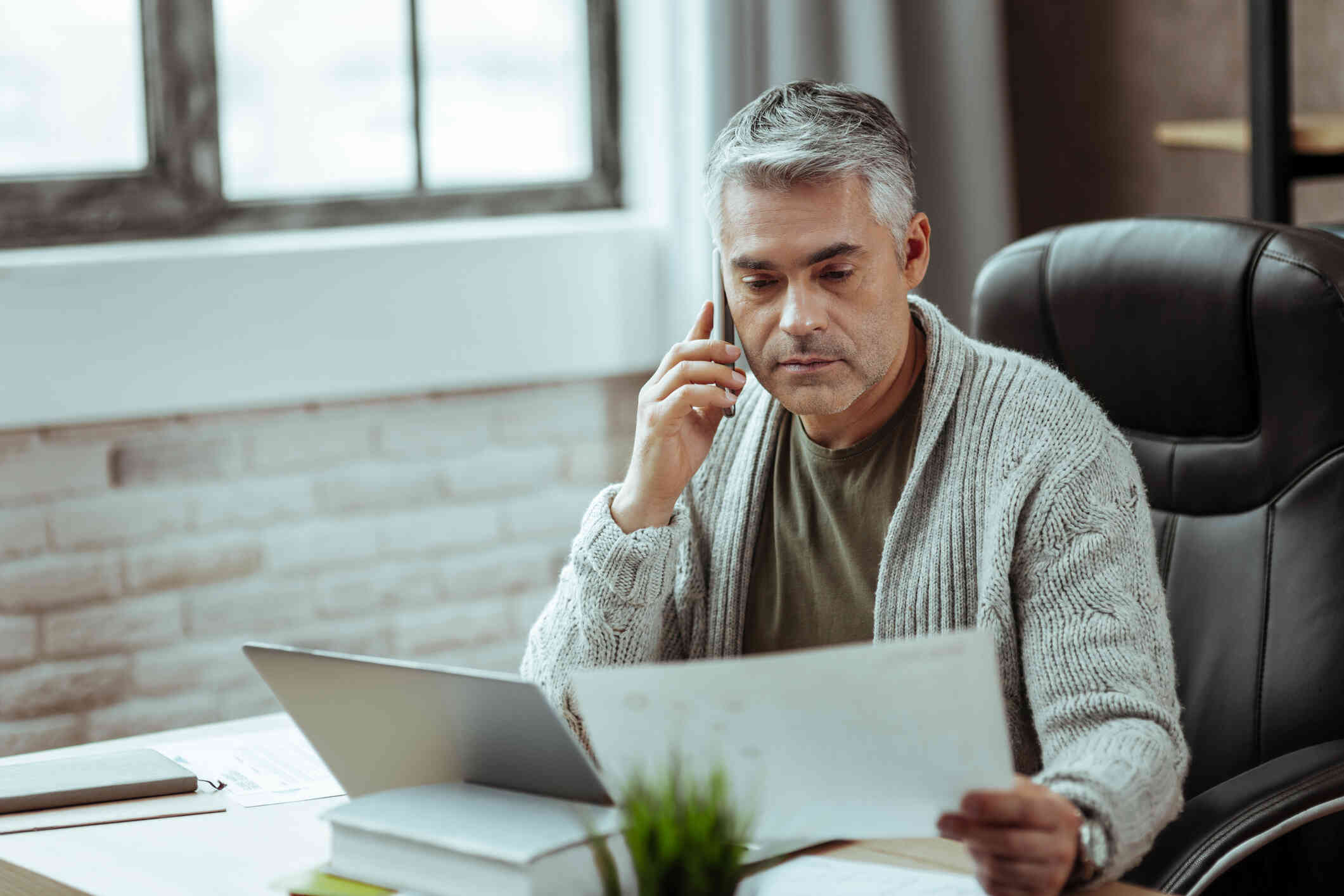 A middle aged man in a grey cardigan sits at his desk and talks on the phone while looking at some papers in his hand.